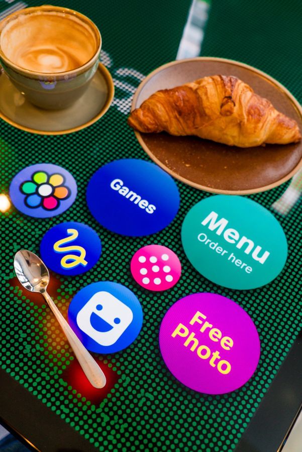 Menu, games and free photo buttons on a Zamarut table, next to a coffee and croissant